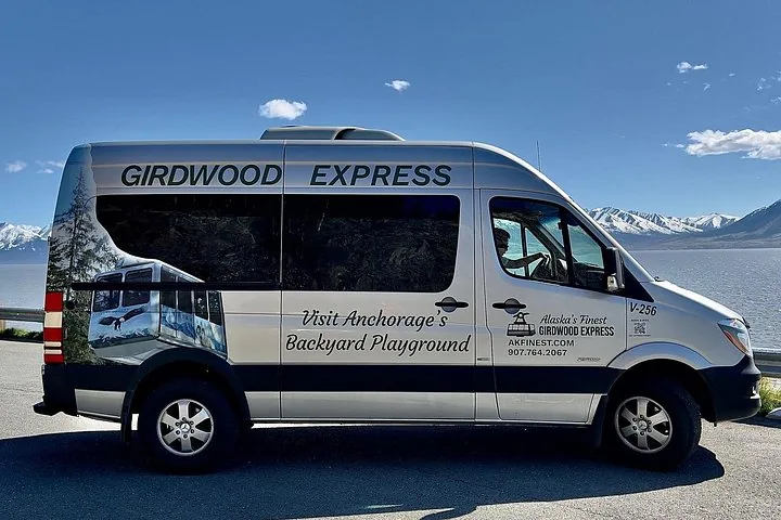 A silver tour van parked near water with a mountainous landscape behind
