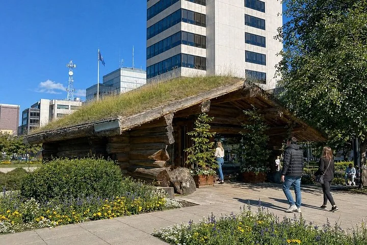 People walking past a wooden cabin with a grass roof in a city