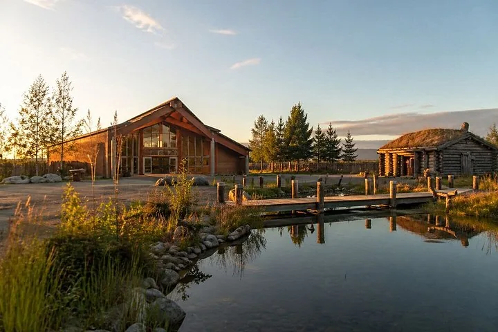 A large wooden building and a log cabin near a calm pond